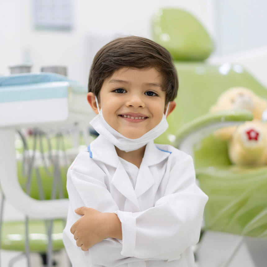A young child dressed in a white lab coat with a stethoscope, giving a thumbs up in a playful imitation of a medical professional, possibly in a pediatric dental clinic setting.